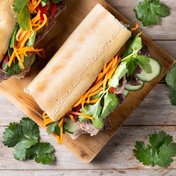 Two Vietnamese banh mi sandwiches with meat, carrots, cucumbers, and cilantro on a wooden board.