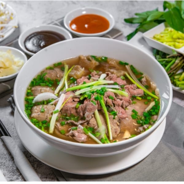 A large bowl of Pho Delight's Vietnamese pho with beef, green onions, and side sauces.