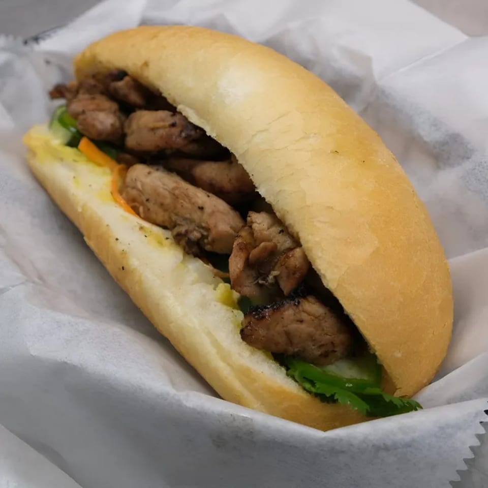 A Vietnamese banh mi sandwich with grilled chicken, pickled vegetables, and cilantro in a white paper wrapper.