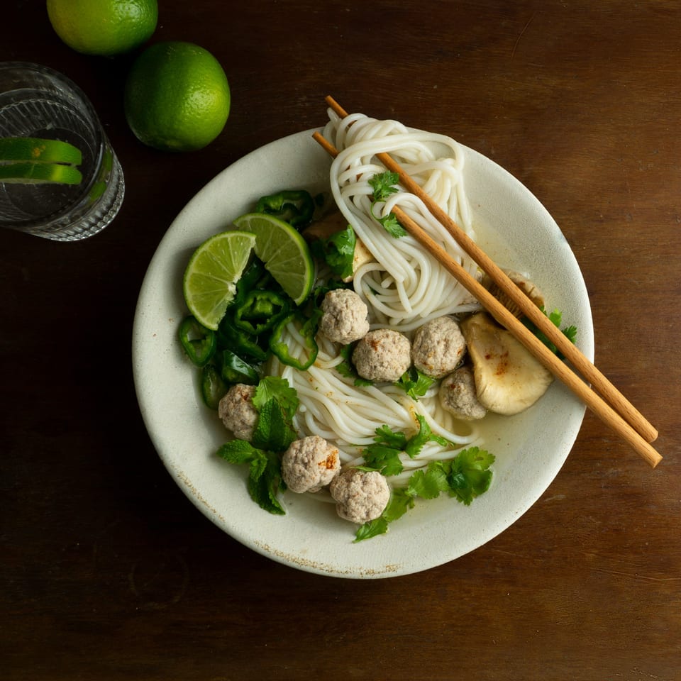 A bowl of Pho Delight's Vietnamese pho with meatballs, noodles, lime, and cilantro, with chopsticks.